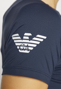 Marineblauw sportoverhemd met korte mouwen van een gestructureerde stof, met een groot wit gestreept adelaarlogo op de bovenste linkerschouder.