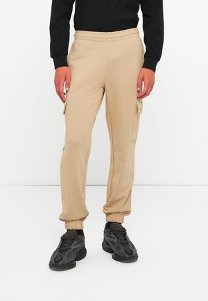 Mand iført beige cargo joggerbukser med elastiske manchetter og sorte chunky sneakers, stående på et hvidt gulv mod en hvid baggrund.