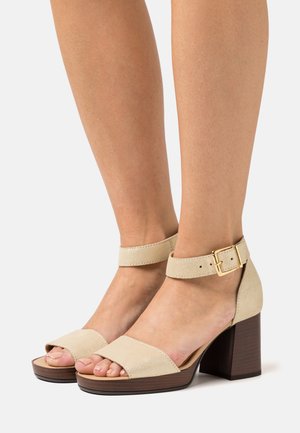 Beige suede block-heel sandals with ankle strap and gold buckle detail. Open-toe design and wooden-like textured heel.