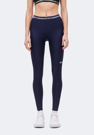 Γυναίκα που φοράει σκούρο μπλε κολάν Under Armour, μαύρο αθλητικό σουτιέν, λευκές κάλτσες και λευκά αθλητικά παπούτσια, στέκεται μπροστά από ένα απλό φόντο.
