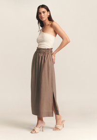 Top de canalé beige claro con un solo hombro, combinado con una falda marrón de cintura alta y fluida que presenta aberturas laterales y una cintura elástica.