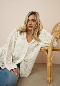 Öppenstickad, vit cardigan med ett geometriskt mönster, med en knappframdel och avslappnade ärmar, kombinerad med ljusa jeans.
