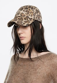 Jonge vrouw met lang, donker haar die een bruine gebreide trui en een luipaardprintmuts draagt, en naar beneden kijkt tegen een witte achtergrond.