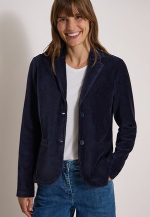 Femme souriante portant un blazer en velours côtelé bleu foncé, une chemise blanche et un jean bleu, une main dans la poche.