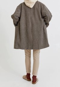 Cappotto oversize a quadri marrone e crema con cappuccio, pantaloni beige e scarpe brown slip-on; texture sottile e silhouette rilassata.