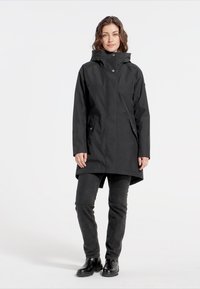 Svart vattentät kappa med hög krage, tryckknappar och fickdetaljer. Modell iklädd mörka byxor och stövlar, ståendes framför en neutral bakgrund.