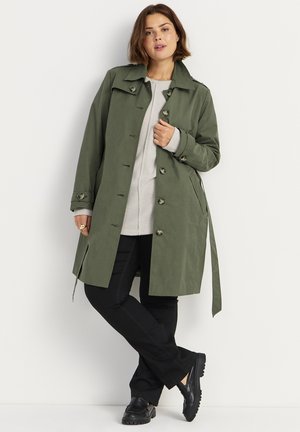Olijfgroene trenchcoat met een op maat gemaakte uitstraling, knoopsluitingen, zijzakken en een strikceintuur, gecombineerd met een lichtgrijze trui en zwarte broek.