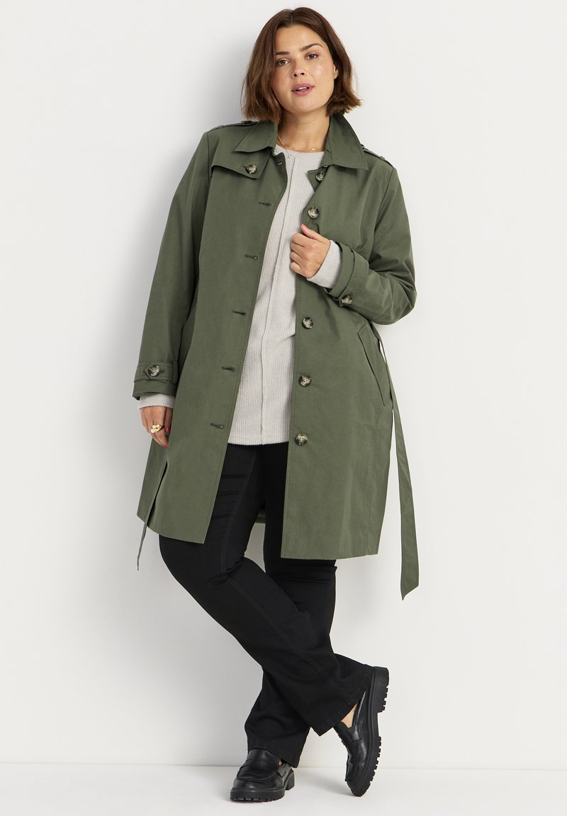 Olijfgroene trenchcoat met een op maat gemaakte uitstraling, knoopsluitingen, zijzakken en een strikceintuur, gecombineerd met een lichtgrijze trui en zwarte broek.