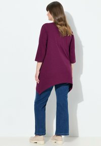 Langärmlige, tiefpurpurfarbene Tunika mit asymmetrischem Saum, kombiniert mit blauen geraden Jeans und beigen Schuhen. Minimalistisches Design und weiche Textur.