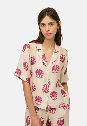 Mujer con camisa beige de manga corta abotonada y pantalones a juego con estampados de hojas rojas, de pie con una mano en el bolsillo.