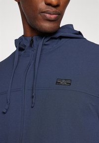 Veste à capuche marine en tissu texturé, dotée d'une fermeture éclair à l'avant et de cordons de serrage réglables. Petite étiquette de marque noire sur la poitrine.