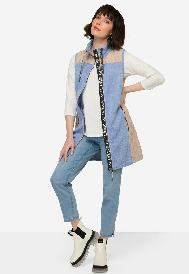 Gilet lungo con frontale blu, retro beige trapuntato e cerniera nera con nastro logo. Abbinato a denim chiaro e stivali alla caviglia bianchi con dettagli neri.