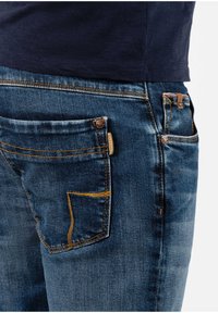 Rückansicht einer Person, die blaue Jeans mit orangefarbenen Nähten und ein in die Hose gestecktes dunkelblaues Hemd trägt.