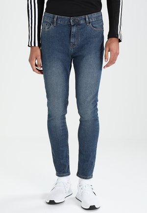 Man draagt een strakke blauwe spijkerbroek, witte sneakers en een zwart shirt met lange mouwen met witte strepen op de mouwen.