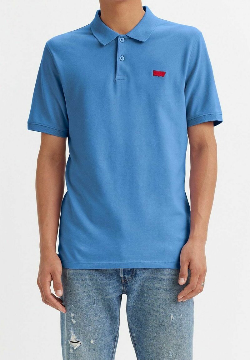 Personne portant un polo bleu uni à manches courtes avec un petit logo rouge et un jean bleu clair usé, debout devant un fond blanc.