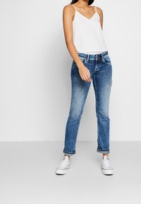 Bílý tílko s tenkými ramínky, spárované s světle modrými džíny s rovnými nohavicemi a ohrnutými manžetami, nošené s bílými teniskami.