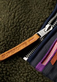 Nahaufnahme einer Fleecejacke, grüner strukturierter Stoff mit mehreren Reißverschlüssen in Marineblau, Lila und Pink. Brauner Lederreißverschlussanhänger mit eingeprägtem Schriftzug "SUPERDRY".