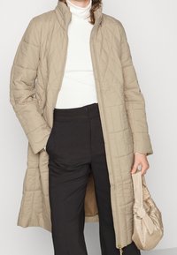 Abrigo largo acolchado beige con cuello alto y cierre de cremallera. Se lleva sobre un suéter de cuello alto blanco y pantalones negros, combinado con un bolso beige.