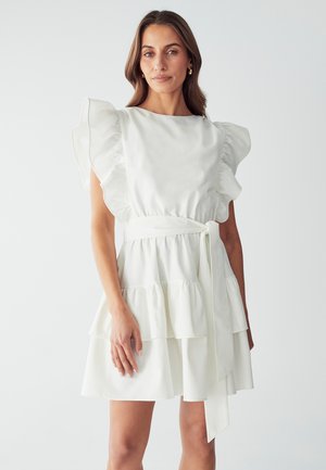 Valkoinen mekko, jossa on röyhelöshortit hihat, kerroksellinen hame ja vyötärösolki. Pehmeä, teksturoitu kangas, jossa on virtaava muotoilu. Yksinkertainen ja elegantti siluetti.