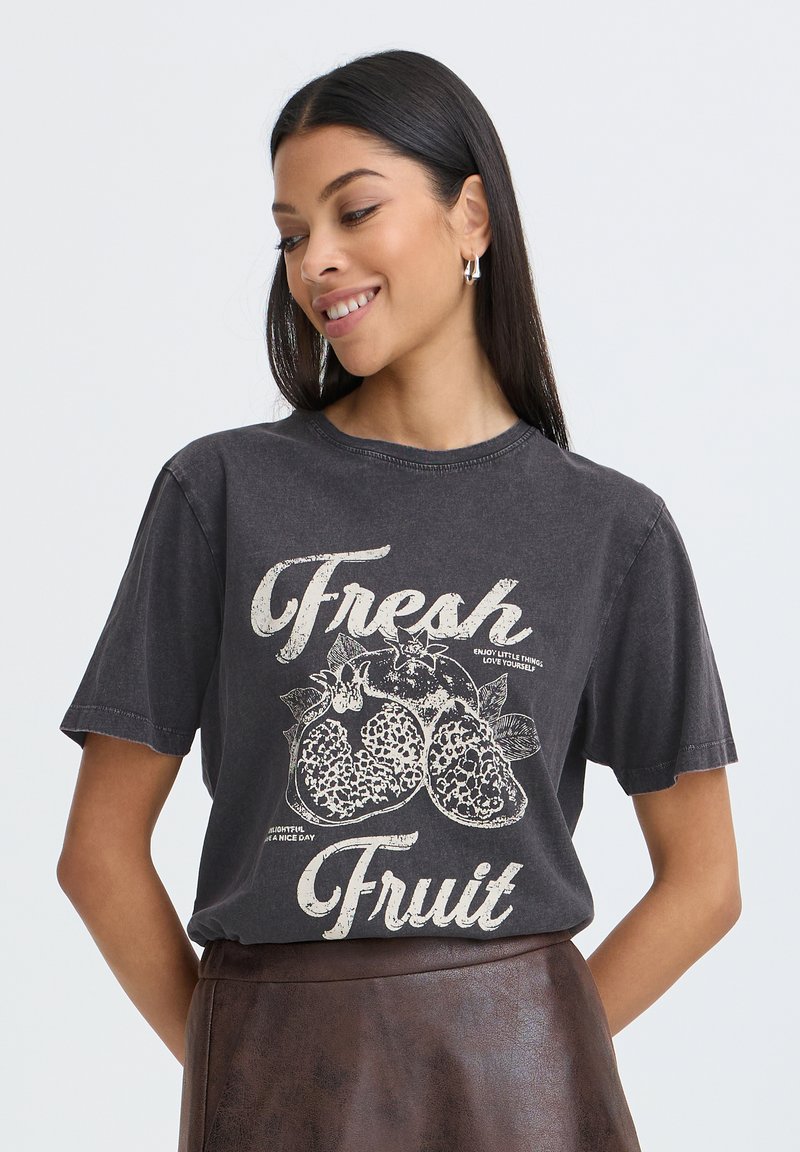 Femme aux longs cheveux foncés portant des boucles d'oreilles créoles argentées et un t-shirt noir avec l'inscription « Fresh Fruit » et des motifs de grenade, associé à une jupe marron.
