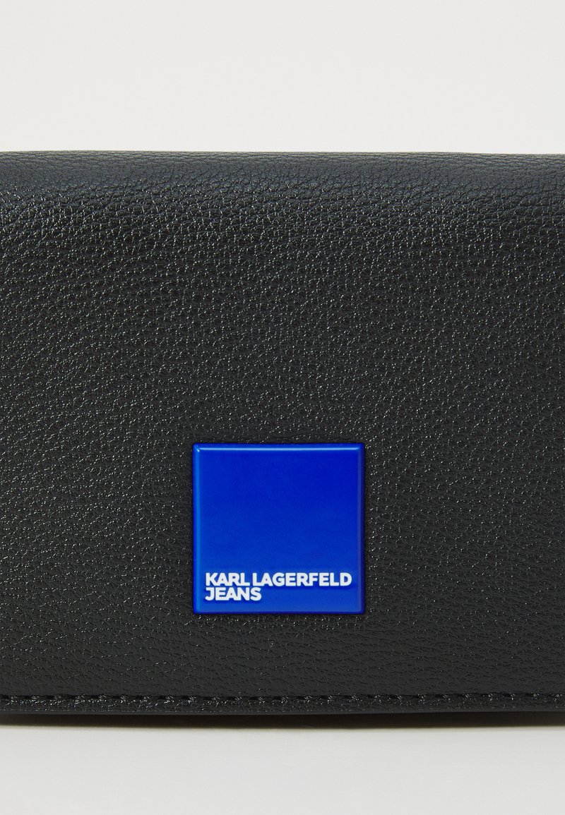 Portefeuille noir texturé avec une étiquette rectangulaire bleue lisse affichant "KARL LAGERFELD JEANS" en blanc, présentant un design minimaliste.