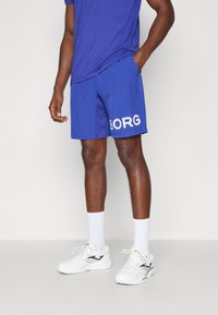 Calções desportivos azuis com o texto branco "BORG" no lado esquerdo, feitos de tecido suave. Usados com meias brancas e ténis, combinados com uma camisa azul.