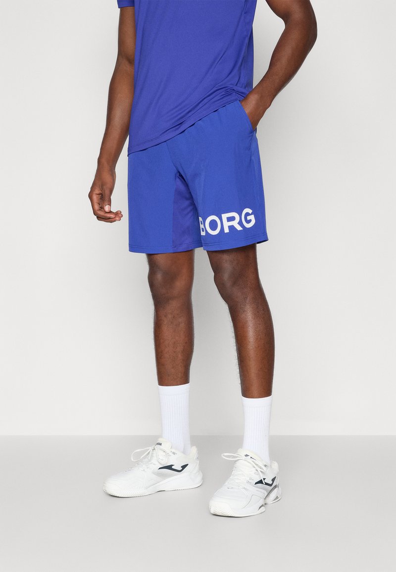 Calções desportivos azuis com o texto branco "BORG" no lado esquerdo, feitos de tecido suave. Usados com meias brancas e ténis, combinados com uma camisa azul.