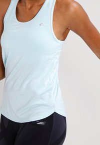 Jasnoniebieski sportowy top na ramiączkach z cienkimi poziomymi paskami, okrągłym dekoltem i zaokrąglonym dołem. Noszony z czarnymi legginsami z logo.