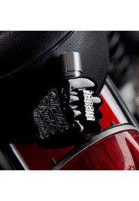 Bouteille de parfum en forme de poing noir avec un capuchon argenté et le logo "DIESEL" en relief. Typographie ornée blanche sur la bouteille, contrastant avec sa finition brillante.