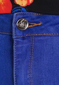 Blå denimjeans med orange sömmar, utrustade med en cirkulär metallknapp med graverad logotyp, texturerat tyg och en synlig midjeresår.