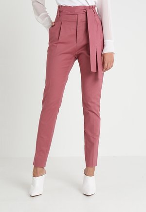 Kvinde iført højtaljede, rosa bukser med bælte og smalle ben, kombineret med en hvid bluse og hvide spidse sko.