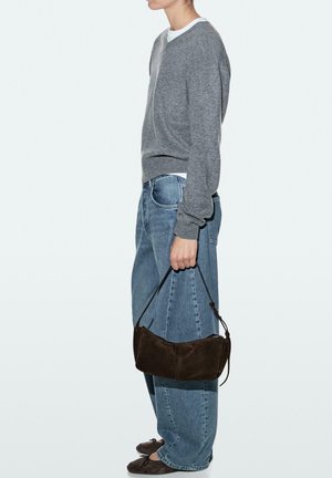 Person in grauem Pullover und blauer Jeans, die eine kleine dunkelbraune Wildlederhandtasche an ihrem Henkel hält, steht vor einem schlichten hellen Hintergrund.