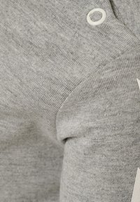 Grå bomullstyg med en texturerad yta, med detaljer på axeln i form av en knapp och ett kontrasterande vitt grafiskt inslag.