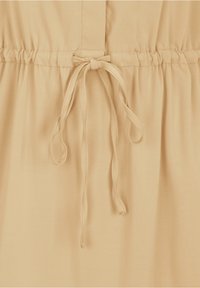 Robe beige présentant une taille froncée avec un lien à nouer, fabriquée en tissu doux, avec une texture lisse et un design minimaliste.