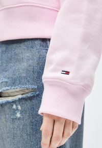 Χέρι που ακουμπά στο πλευρό, φοράει ανοιχτό ροζ φούτερ με μικρό λογότυπο στη μανσέτα και σκισμένο στενό τζιν μπλε.