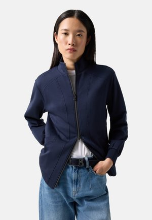 Frau trägt eine dunkelblaue Jacke mit Reißverschluss über einem weißen Hemd und lockere blaue Jeans mit schwarzem Gürtel, steht mit einer Hand in der Tasche.