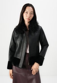 Veste en cuir noir avec col et poignets en fausse fourrure, dotée d'une fermeture éclair à l'avant et d'une texture unie, associée à une jupe en cuir bordeaux.