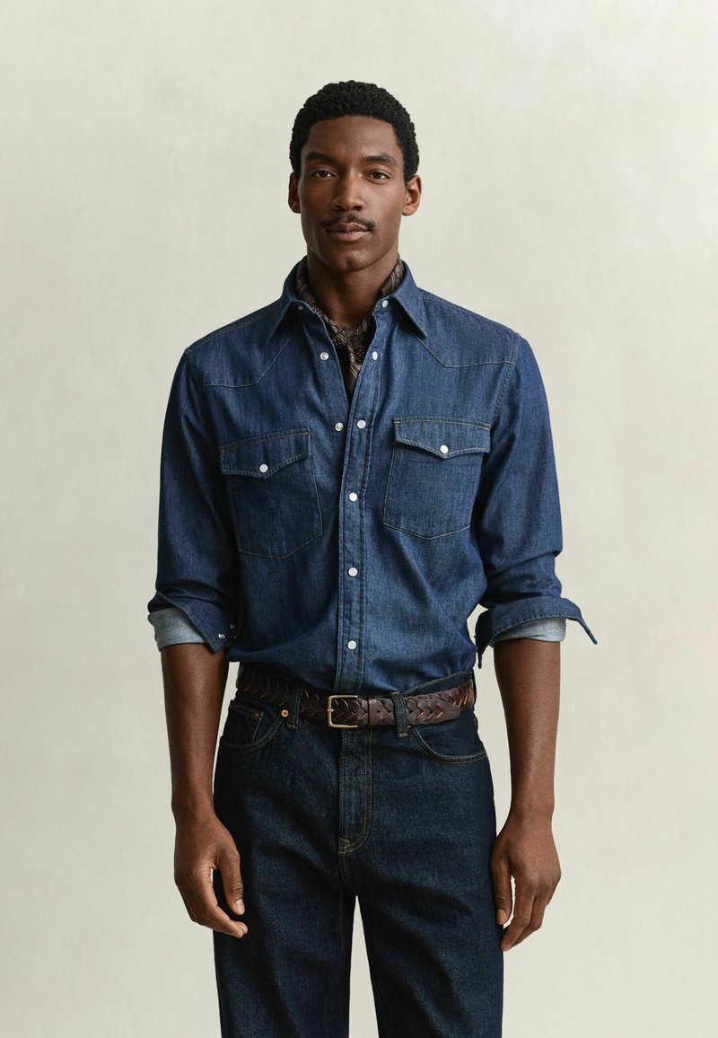 Man with short curly hair and mustache wears dark denim shirt with rolled sleeves, dark jeans, and braided leather belt against plain background.