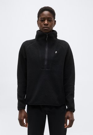 Osoba ubrana w czarny polar z wysokim kołnierzem i zamkiem do połowy, stojąca na jednolitym jasnoszarym tle.