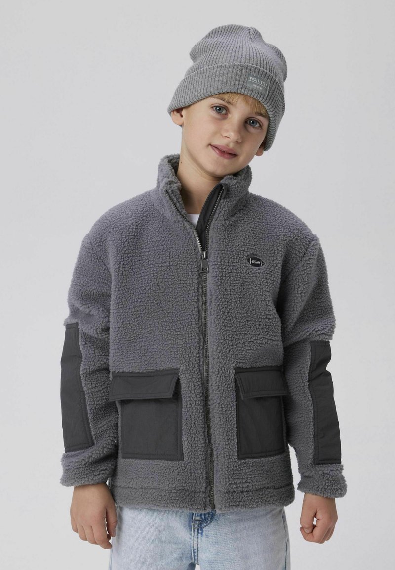 Harmaa fleece-takki mustilla kankaan yksityiskohdilla ja taskuilla. Vetoketjukiinnitys, korkea kaulus ja ribbattu harmaa pipo. Rentoa muotoilua.