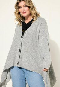 Cardigan grigio chunky con scollo a V, due bottoni frontali e orlo asimmetrico. Texture morbida, vestibilità rilassata sopra una T-shirt nera e jeans con decorazioni.