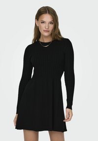 Robe noire à manches longues côtelées avec un corsage ajusté et une jupe évasée, fabriquée en matière douce. Collier doré en accessoire.