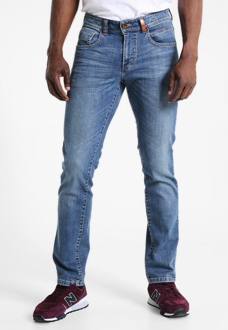 Homme portant un jean slim bleu clair et des baskets New Balance bordeaux, debout devant un fond blanc.