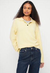 Pull-over en tricot jaune avec un col à revers, deux boutons noirs, et des poignets à côtes, assorti à un jean bleu foncé.