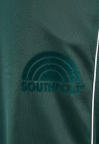 Zöld textil, amelyen egy kiemelkedő sötétzöld bársony "SOUTHPOL" logó látható, szivárvány mintával és fehér pántos díszítéssel.