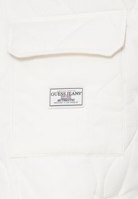 Bijela kviltana tkanina s prednjim džepom i preklopom. Sadrži oznaku koja glasi "GUESS JEANS", s detaljima o datumu osnivanja i mjestu dizajna.