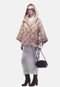 Beige gewatteerde poncho met capuchon en een losse fit, gecombineerd met een lange lichtgrijze rok en bruine hooghakken. Gecombineerd met een kleine donkere tas.