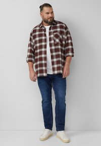 Flanellen overhemd in bordeauxrood en witte ruit met knoopsluiting aan de voorkant en een borstzak, gecombineerd met donkerblauwe spijkerbroek en witte sneakers.