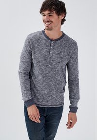 Maglietta henley a maniche lunghe in grigio scuro fruttato con quattro bottoni decorativi, polsi a coste e scollatura rotonda. Indossata con jeans scuri.