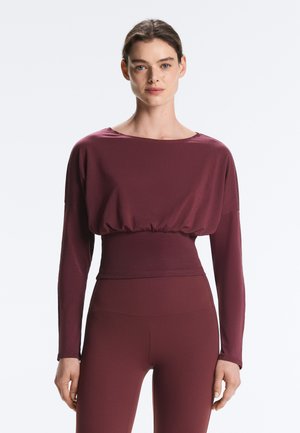 Žena stojaca, oblečená v priliehavom, dlhom rukáve vínovo sfarbeného topu so zúženým pásom a rovnakých vínovo sfarbených legín na bielom pozadí.